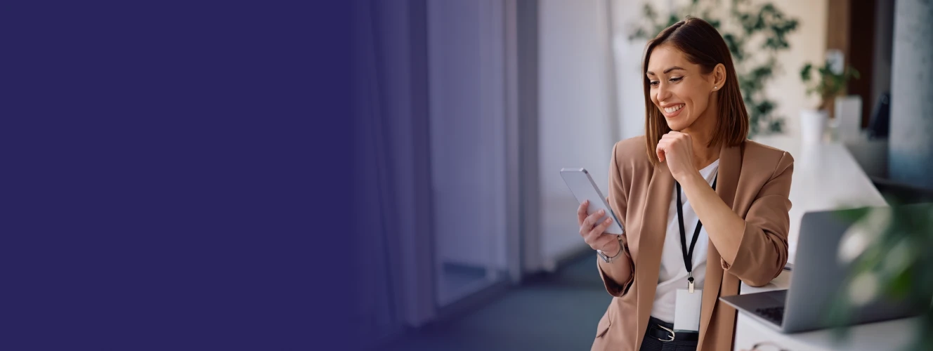 Directrice d’usine souriante consultant les modules ERP sur son smartphone, debout près d’un bureau avec un ordinateur portable.